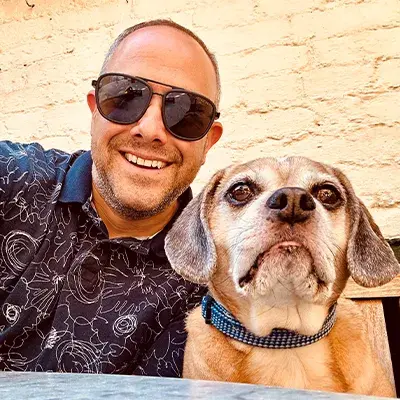 A man smiling wearing sun glasses and posing with a dog that is looking at the camera